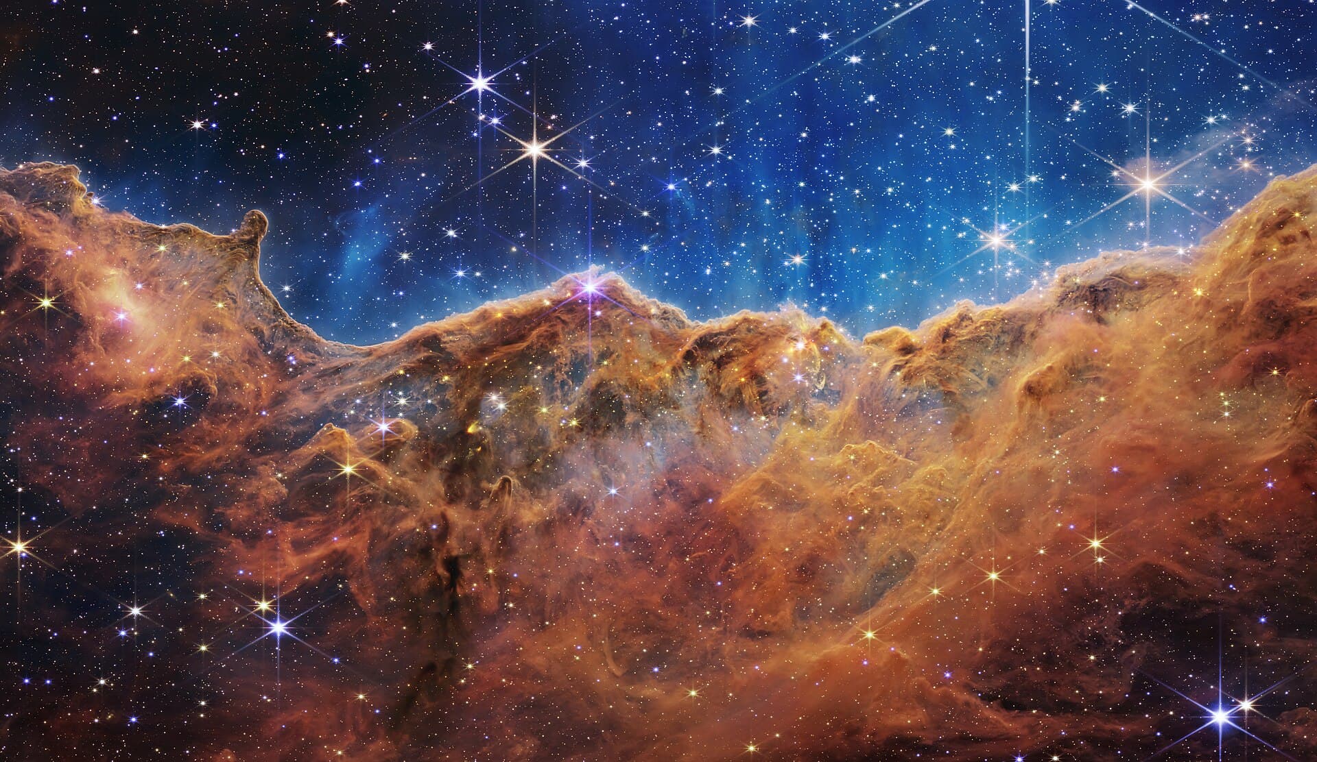 Towering clouds of gas and dust glowing orange and gold against a deep blue sky in the Carina Nebula